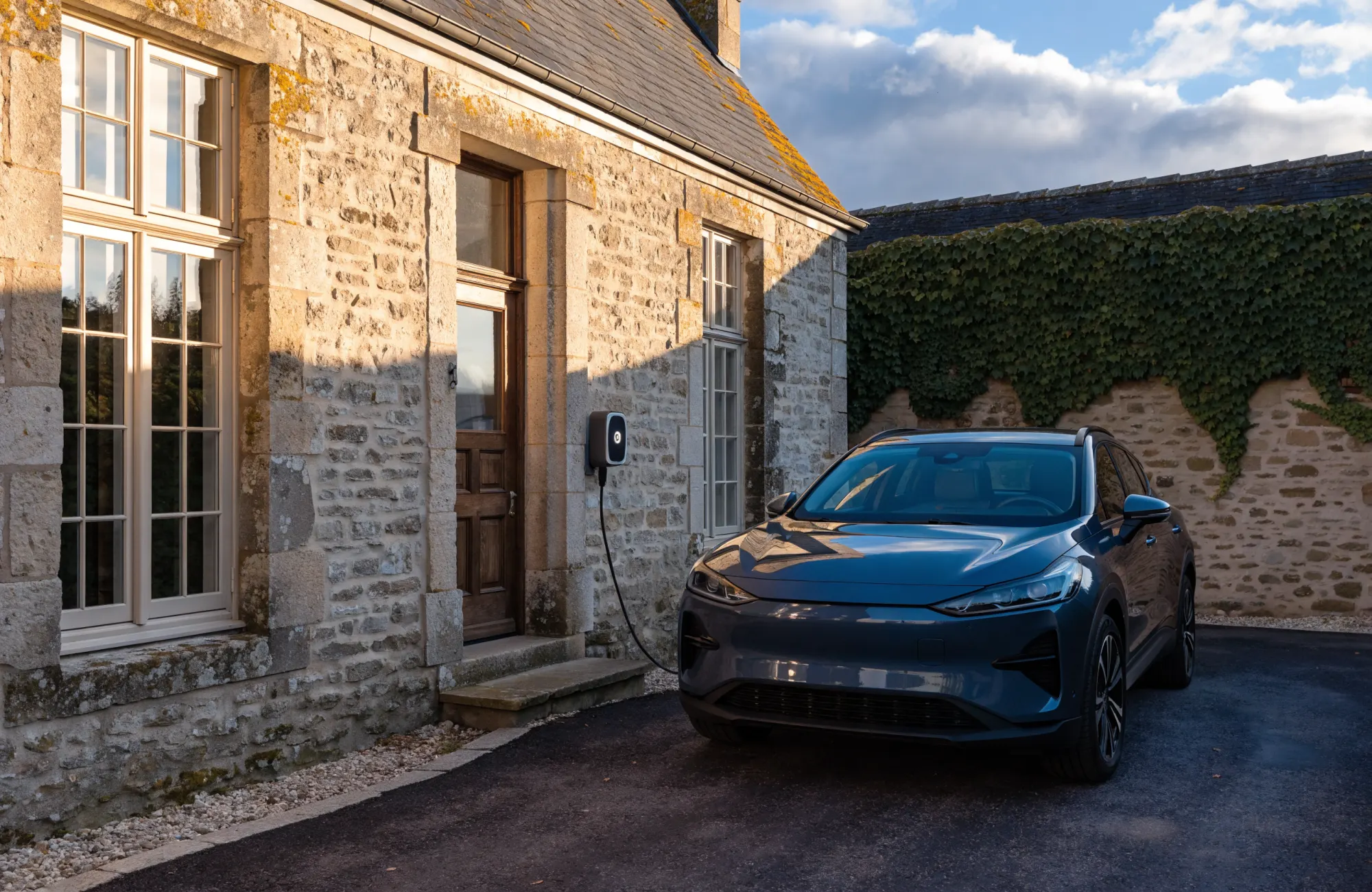 Borne de recharge murale posée sur la façade en pierre d’une maison, une voiture électrique branchée dans l’allée