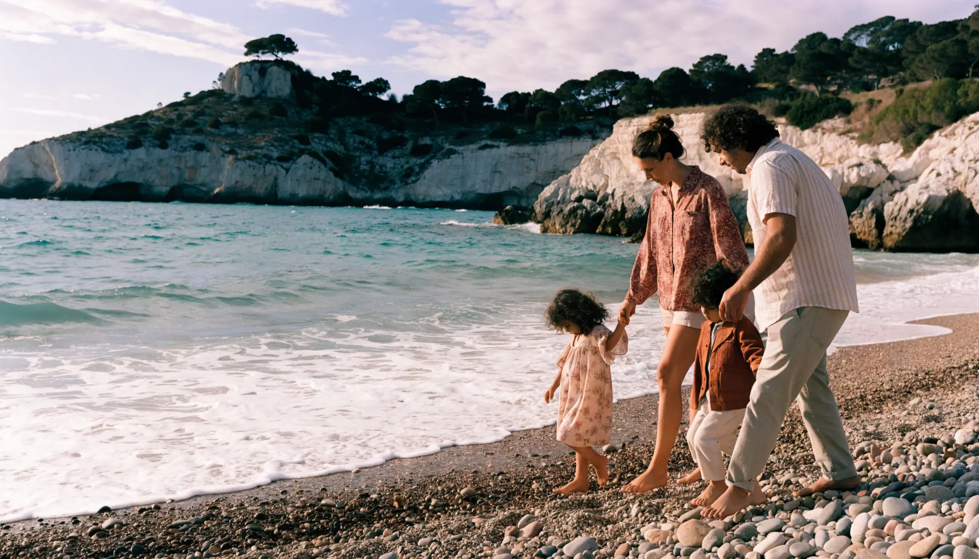 Une famille de quatre marche pieds nus sur une plage de galets dans une calanque, au coucher du soleil